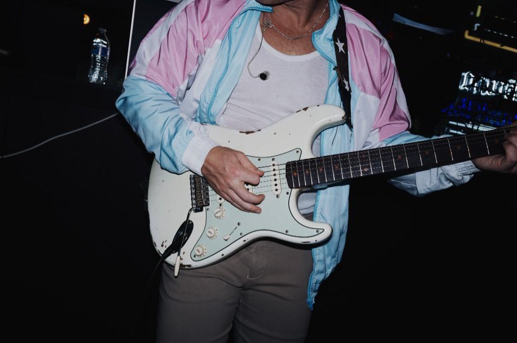 Luke Lively playing a white electric guitar on stage in a light blue and pink windbreaker